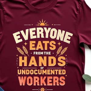May include: Maroon t-shirt with the text "EVERYONE EATS from the HANDS of UNDOCUMENTED WORKERS" in white and orange. The shirt is paired with blue jeans and a tan hat. A sprig of green leaves is also in the image.