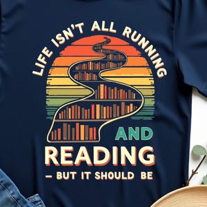 Può includere: T-shirt blu navy con un design grafico raffigurante un tramonto, un percorso tortuoso di librerie e la scritta "LIFE ISN'T ALL RUNNING AND READING - BUT IT SHOULD BE". La maglietta è abbinata a jeans blu e un cappello di paglia.