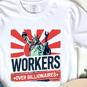 May include: White t-shirt with a graphic of the Statue of Liberty holding a torch and wrench, set against a red and white background. The text "WORKERS OVER BILLIONAIRES" is displayed below the image. Blue jeans and a beige hat are also in the image.
