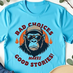 Może przedstawiać: Jasnoniebieska koszulka z grafiką goryla w okularach przeciwsłonecznych i czapce, z napisem "BAD CHOICES MAKES GOOD STORIES!". Na zdjęciu widoczne są również niebieskie dżinsy i beżowy kapelusz.