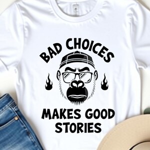 Może przedstawiać: Biała koszulka z napisem "BAD CHOICES MAKES GOOD STORIES" i grafiką goryla w okularach i kapeluszu. Koszulka jest zestawiona z niebieskimi dżinsami i beżowym kapeluszem.