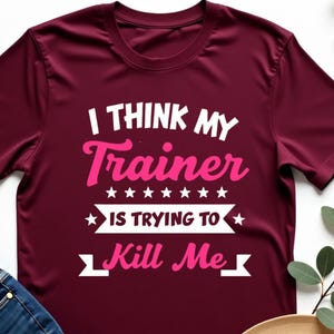 Pode incluir: Camiseta bordô com o texto "I THINK MY Trainer IS TRYING TO Kill Me" em branco e rosa. A camisa é combinada com jeans azuis e um chapéu bege.