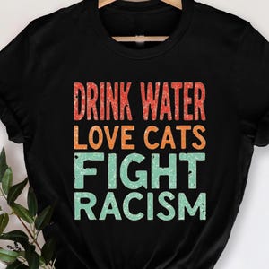 This funny Cat Mom t-shirt is perfect for cat lovers who are passionate about animals and standing up against racism. Featuring a witty message that combines a love for cats with a call to fight racism, this shirt makes a bold statement. The comfortable design is perfect for everyday wear, while the message speaks to the values of kindness, activism, and self-care. Drink water, love cats, and fight for equality with this unique, eye-catching tee that every cat lover will adore.