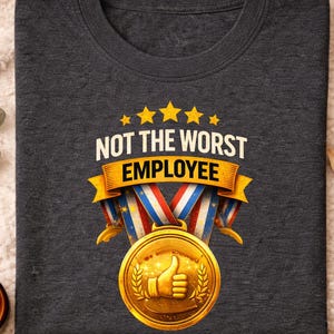 May include: Dark gray heather crew neck t-shirt with the text "NOT THE WORST EMPLOYEE" above a gold medal with a thumbs-up. The medal has red, white, and blue ribbons and is surrounded by a laurel wreath. The shirt is on a soft, textured surface.