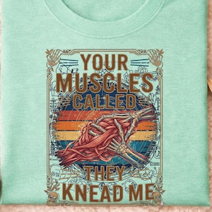 Op de afbeelding: Mintgroen T-shirt met ronde hals en een grafisch ontwerp. Het ontwerp toont een gestileerde illustratie van spieren en botten in een cirkelvormig kader, met de tekst "YOUR MUSCLES CALLED THEY KNEAD ME."