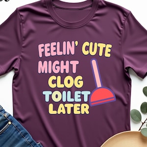 May include: A maroon t-shirt with the text "Feelin' Cute Might Clog Toilet Later" in colorful letters. A cartoon plunger is next to the text.