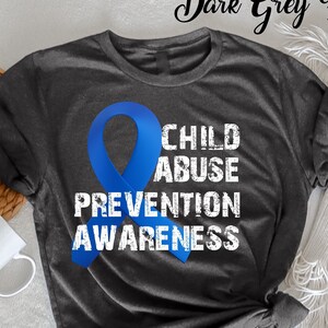 Könnte beinhalten: Dunkelgraues Heather-T-Shirt mit blauem Band und dem weißen Schriftzug „CHILD ABUSE PREVENTION AWARENESS“. Das Shirt liegt gefaltet auf einer weißen Oberfläche, mit einem geflochtenen Korb und einer weißen Tasse.