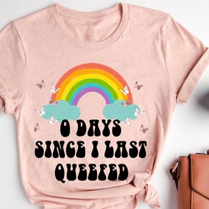 May include: A pink t-shirt with a rainbow and clouds graphic. The text on the shirt reads "0 days since I last queefed".