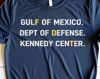 Könnte beinhalten: Ein marineblaues T-Shirt mit weißem Text: "GULF OF MEXICO. DEPT OF DEFENSE. KENNEDY CENTER." Die Buchstaben "F", "D" und "T" sind gelb. Die Worte "Heather Navy" sind in Kursivschrift oben rechts geschrieben.
