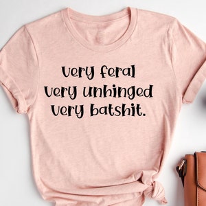 May include: A pink t-shirt with the text "Very feral very unhinged very batshit." printed on it.