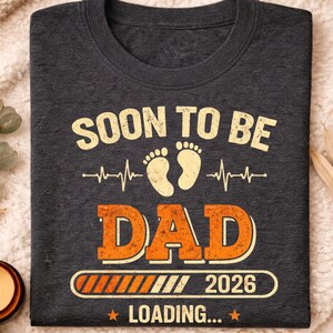 May include: Dark grey heather crew neck t-shirt with the text "SOON TO BE DAD 2026 LOADING..." in orange and cream colours. The design includes baby footprints and a heartbeat line. The shirt is folded on a cream-coloured blanket.