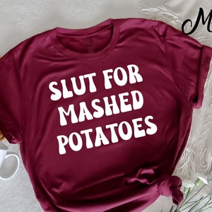 Op de afbeelding: Een maroon t-shirt met de tekst "SLUT FOR MASHED POTATOES" in wit.