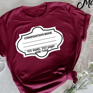 Op de afbeelding: Bordeauxrood T-shirt met een wit labelontwerp met de tekst "COMPOSITION BOOK" en lijnen om op te schrijven. Het label vermeldt ook "100 sheets, 200 pages wide ruled". Het shirt is aan de onderkant geknoopt.
