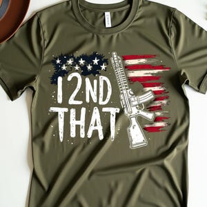 Puede incluir: Camiseta verde oliva con un diseño gráfico que presenta la bandera estadounidense, un rifle y el texto "12ND THAT" en blanco. Un sombrero marrón es visible en la esquina superior izquierda. La camiseta está hecha de un material suave y cómodo.