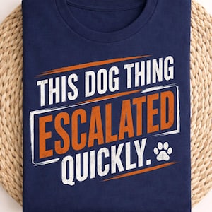 May include: A navy blue t-shirt featuring the white and orange text "THIS DOG THING ESCALATED QUICKLY." with a small white paw print icon. The design is centered on the front of the shirt, utilizing a bold, distressed font style for the lettering.