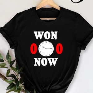 Pode incluir: Camiseta preta com texto branco que diz "WON 0 0 NOW" e um mostrador de relógio com ponteiros vermelhos.