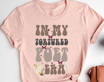 May include: Pale pink t-shirt featuring the text "IN MY TORTURED POET ERA" in a retro brown and grey font. The design includes butterfly graphics and a quill pen illustration. The brand name "Heather Peach" is written in cursive at the top.