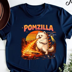Pode incluir: Camiseta azul marinho com um desenho animado de um cão Pomerânia cuspindo fogo, com o texto "POMZILLA". O design inclui um pôr do sol, um horizonte da cidade, um carro e um helicóptero.