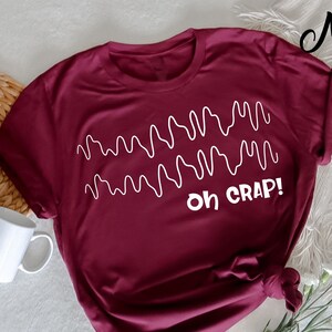 Op de afbeelding: Een bordeauxrood t-shirt met een witte afbeelding van een golvend lijnenpatroon en de tekst "Oh Crap!"