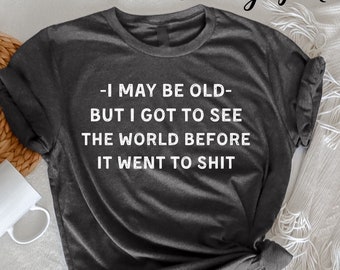 Puede incluir: Camiseta gris oscuro jaspeado con el texto en blanco "-I MAY BE OLD- BUT I GOT TO SEE THE WORLD BEFORE IT WENT TO SHIT". La camiseta se muestra sobre una superficie blanca con una taza blanca y elementos decorativos.