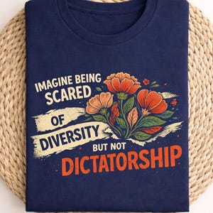 Może przedstawiać: Granatowa koszulka z napisem "IMAGINE BEING SCARED OF DIVERSITY BUT NOT DICTATORSHIP" oraz kwiatowym wzorem w kolorze pomarańczowym i brzoskwiniowym. Koszulka jest złożona na plecionej macie.