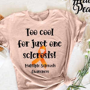 May include: A light brown t-shirt with the text "Too cool for just one sclerosis!" and an orange ribbon. The text "Multiple Sclerosis Awareness" is also printed on the shirt.