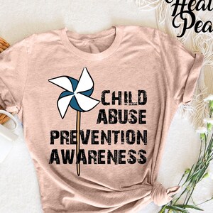 Könnte beinhalten: Hellrosa T-Shirt mit einem Windrad-Motiv und dem schwarzen Text "CHILD ABUSE PREVENTION AWARENESS". Das T-Shirt ist gefaltet, mit einer weißen Tasse und dekorativen Elementen im Hintergrund.