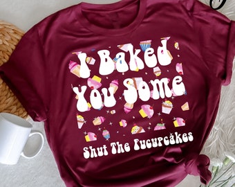 Peut inclure: T-shirt bordeaux avec l'inscription blanche "I Baked You Some Shut The Fucupcakes" et des illustrations de cupcakes. Le t-shirt est de couleur bordeaux unie et a des manches retroussées.