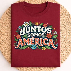 Può includere: T-shirt color bordeaux con la scritta "Juntos Somos America" in un design floreale. Il design include fiori colorati e foglie verdi. La maglietta è piegata su un tappetino intrecciato.