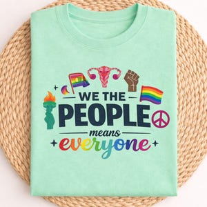Könnte beinhalten: Ein mintgrünes T-Shirt mit dem Text "WE THE PEOPLE means everyone" in dunkler blauer Schrift. Das Design beinhaltet farbenfrohe Grafiken einer Regenbogenflagge, einer Faust, einem Friedenszeichen und einer Fackel.