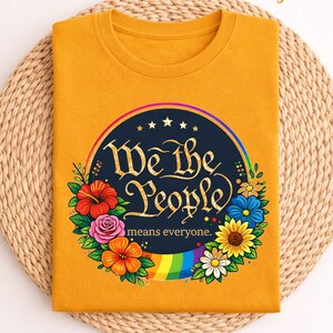 May include: Mustard yellow t-shirt with the text "We the People means everyone" in a circular design. The design includes a rainbow, flowers, and stars. The shirt is folded on a woven mat.