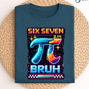 Könnte beinhalten: Türkisfarbenes T-Shirt mit einem pixeligen Grafikdesign des Pi-Symbols in leuchtenden Farben, dem Text "SIX SEVEN 3.14 BRUH" in einer Retro-Schriftart. Das Shirt liegt gefaltet auf einer gewebten, kreisförmigen Matte.