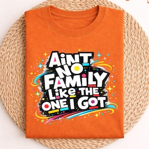 Può includere: T-shirt arancione con la scritta "Ain't No Family Like The One I Got" in un design colorato e stilizzato. La maglietta è piegata e appoggiata su un tappetino rotondo intrecciato.