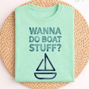 Peut inclure: Un t-shirt vert clair avec le texte "WANNA DO BOAT STUFF?" en bleu sarcelle foncé. En dessous, un dessin de voilier bleu sarcelle. Le t-shirt est présenté sur un tapis rond en osier.