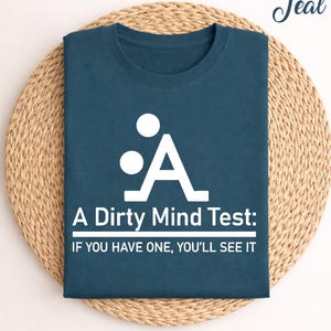May include: Teal-colored t-shirt with a white graphic and text. The graphic is an abstract design with the text "A Dirty Mind Test: IF YOU HAVE ONE, YOU'LL SEE IT". The shirt is folded on a woven mat.