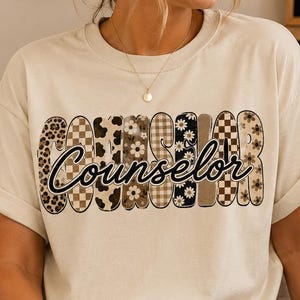 Op de afbeelding: Een crèmekleurig T-shirt met korte mouwen en het woord COUNSELOR in grote, blokvormige letters gevuld met diverse patronen, waaronder luipaardprint, ruitjes en madeliefjes, met het woord Counselor in zwarte schrijfletters eroverheen.