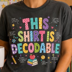 Op de afbeelding: Een antracietgrijs T-shirt met korte mouwen en de kleurrijke tekst "THIS SHIRT IS DECODABLE" met kleine educatieve afbeeldingen en notities, gecombineerd met een witte keramische mok met de tekst "Teach Love Inspire".