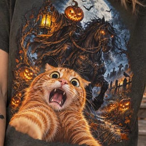 Op de afbeelding: Een antracietgrijs T-shirt met korte mouwen en een grafische print van een oranje cyperse kat met wijd opengesperde ogen op de voorgrond. Achter de kat rijdt een ruiter zonder hoofd op een donker paard onder een volle maan, omringd door pompoenen.