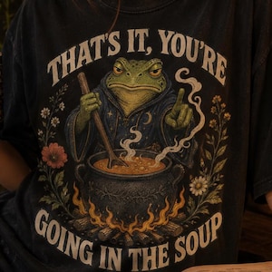 May include: A black short-sleeve t-shirt featuring a graphic of a green frog in a dark robe stirring a steaming cauldron over a fire. The text reads, "THAT'S IT, YOU'RE GOING IN THE SOUP," surrounded by floral illustrations and botanical elements.