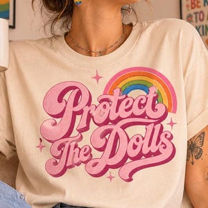 Könnte beinhalten: Ein cremefarbenes T-Shirt mit dem Schriftzug "Protect The Dolls" in einer großen, rosafarbenen Retro-Schriftart. Über dem Text befindet sich eine bunte Regenbogen-Grafik mit kleinen Sternakzenten. Das T-Shirt wird mit einer hellblauen Jeans getragen.