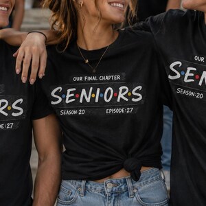 May include: A black short-sleeved t-shirt featuring the text "OUR FINAL CHAPTER" above the word "SENIORS" in a stylized font with colored dots between letters, and "SEASON:20 EPISODE:27" printed below, mimicking a television show title card.
