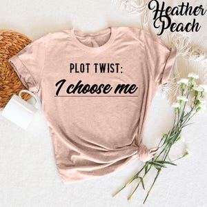 May include: A heather peach colored t-shirt with the text "PLOT TWIST: I choose me" printed in black.