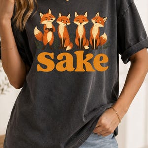 May include: A charcoal gray short-sleeve t-shirt featuring a graphic print of four orange foxes standing in a row above the word "Sake" in large, bold, orange, retro-style lettering. The shirt has a crew neckline and a relaxed, vintage-washed finish.