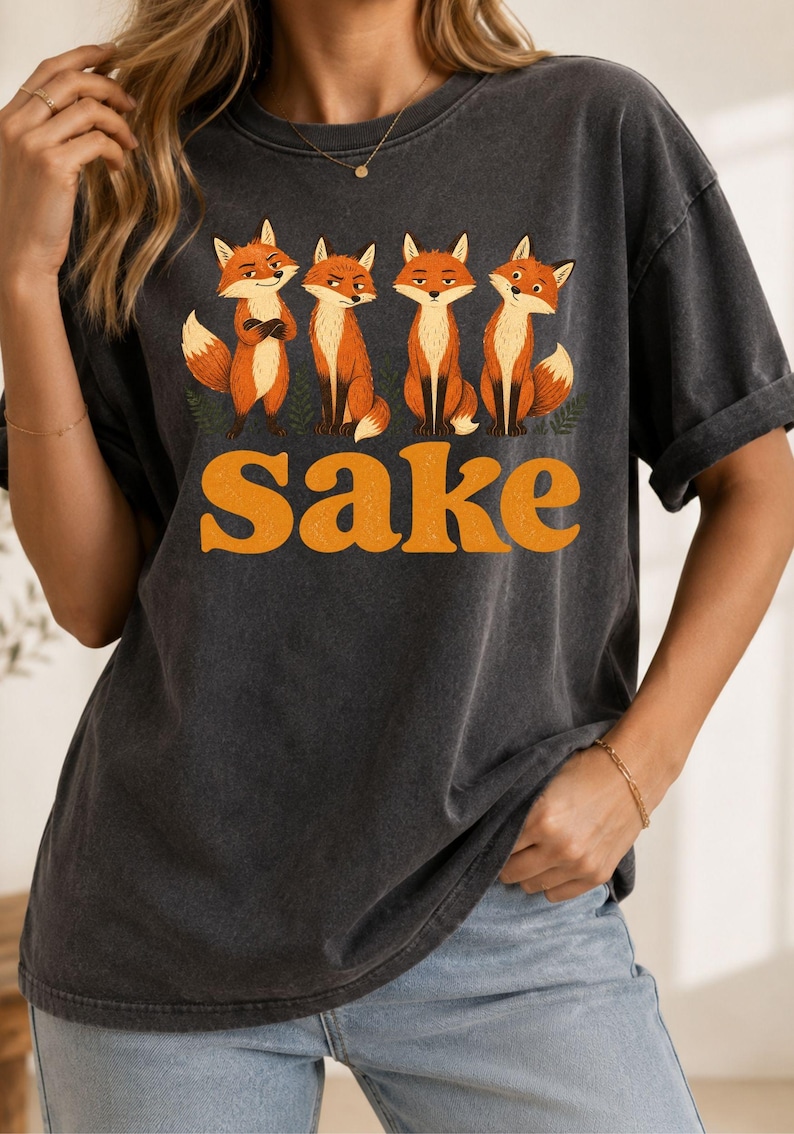 May include: A charcoal gray short-sleeve t-shirt featuring a graphic print of four orange foxes standing in a row above the word "Sake" in large, bold, orange, retro-style lettering. The shirt has a crew neckline and a relaxed, vintage-washed finish.
