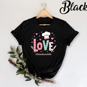 Pode incluir: Camiseta preta com um design gr&aacute;fico da palavra "LOVE" em rosa, verde e branco com um chap&eacute;u de cozinheiro e cora&ccedil;&otilde;es. O texto "#SchoolLunchlife" est&aacute; abaixo do gr&aacute;fico.