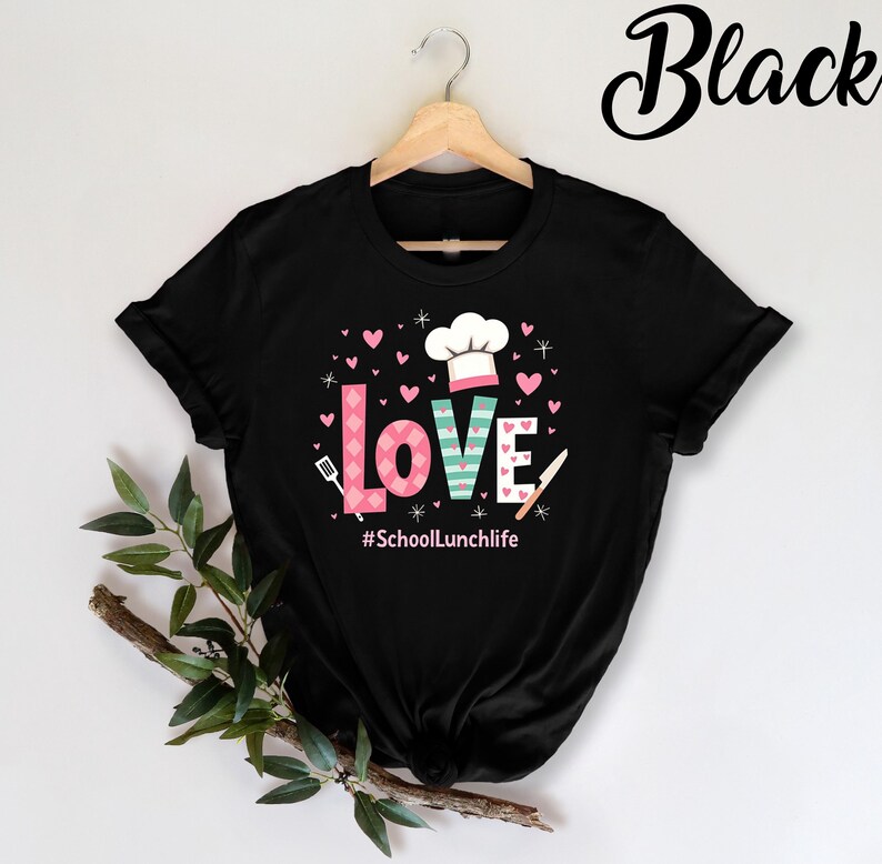 Pode incluir: Camiseta preta com um design gr&aacute;fico da palavra "LOVE" em rosa, verde e branco com um chap&eacute;u de cozinheiro e cora&ccedil;&otilde;es. O texto "#SchoolLunchlife" est&aacute; abaixo do gr&aacute;fico.