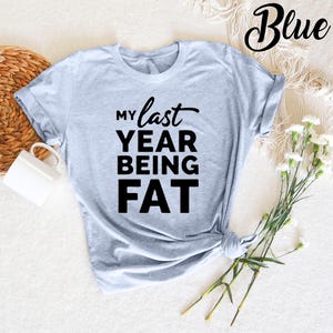 May include: A light blue t-shirt with the text "My last year being fat" printed in black.