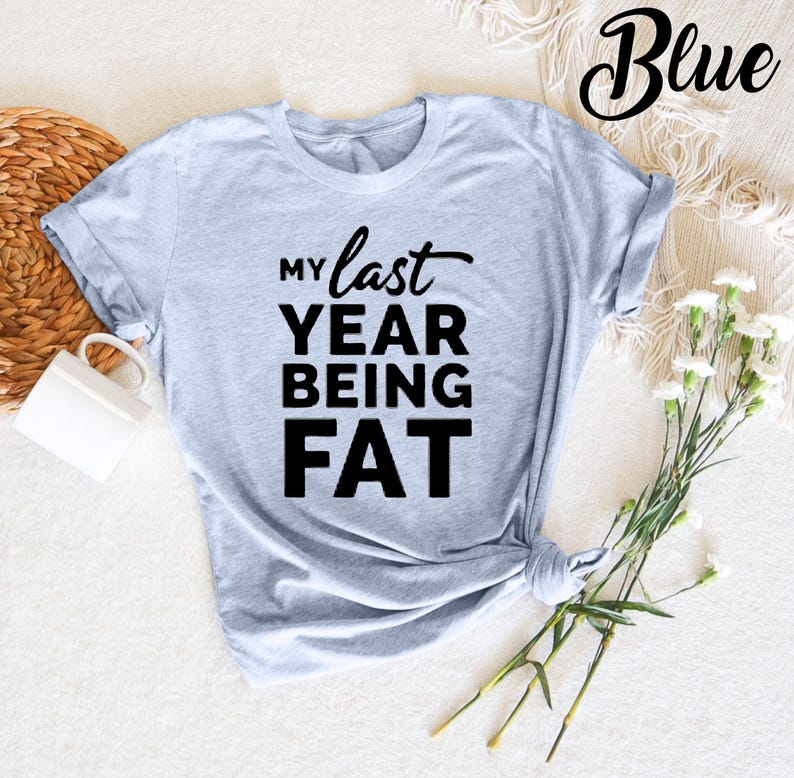 May include: A light blue t-shirt with the text "My last year being fat" printed in black.