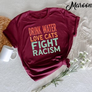 May include: A maroon t-shirt with the text "Drink Water Love Cats Fight Racism" in a retro style font.