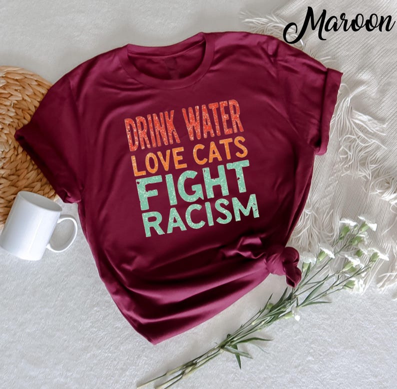 May include: A maroon t-shirt with the text "Drink Water Love Cats Fight Racism" in a retro style font.
